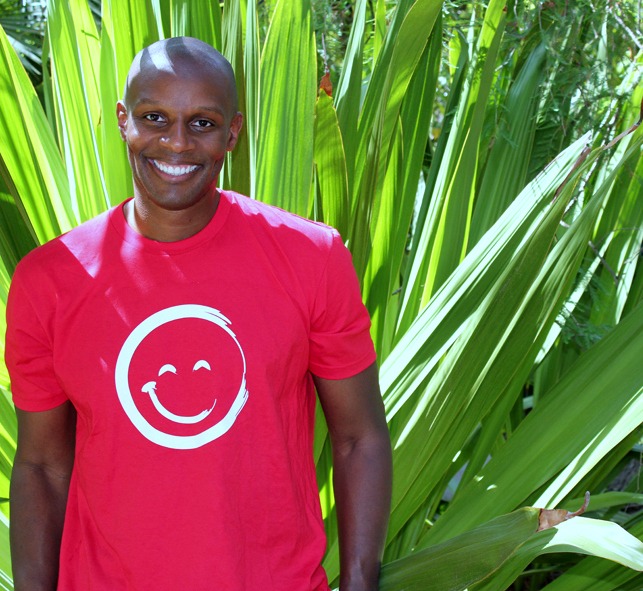 Richard Shola Smiling And Wearing a Red Smiley Face T-Shirt While Standing In Front of Large Plant