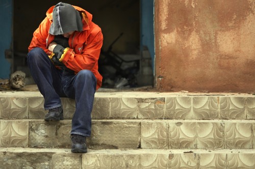 Man in Hoodie With Head Bowed Down Sitting on Top of Steps In Front of Abandoned Building