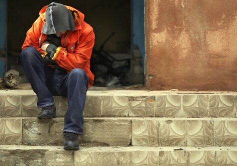 Man in Hoodie With Head Bowed Down Sitting on Top of Steps In Front of Abandoned Building